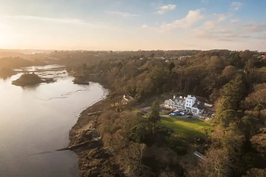 Aerial view of a grand white country house nestled among woodland on a tidal estuary at golden hour, Cornwall