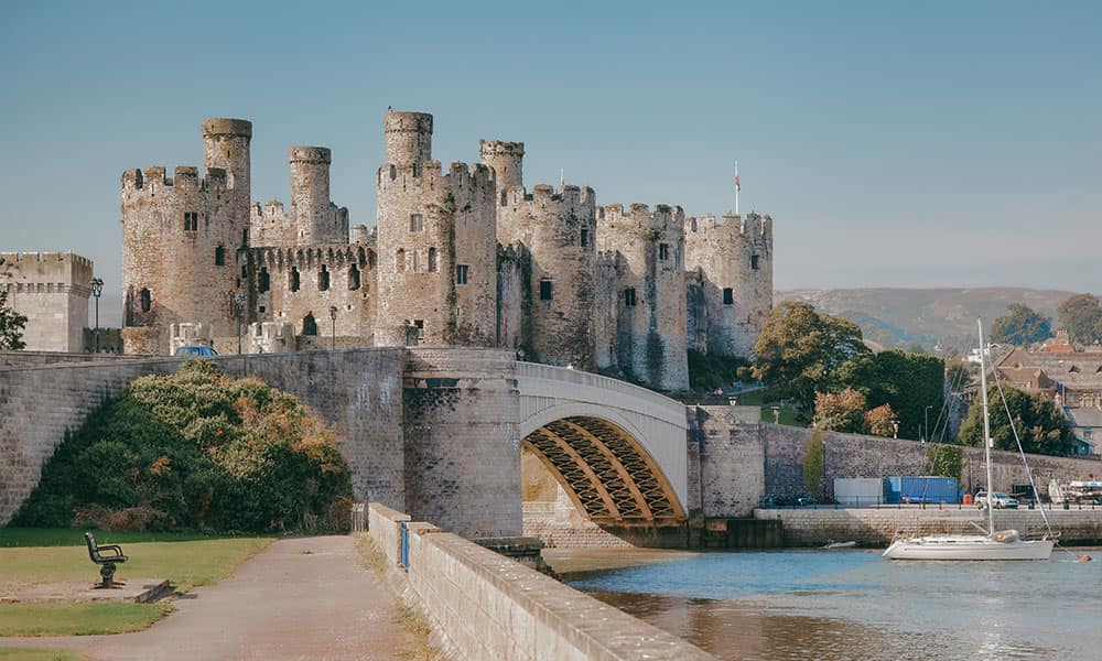 Conwy Castle's medieval towers rising above the River Conwy, with a stone arch bridge and moored sailboat on a sunny day in…