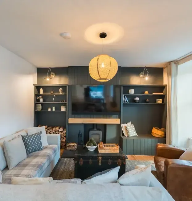 Cosy holiday cottage living room with dark-painted built-in shelving, log burner, wall-mounted TV, and rattan pendant light…
