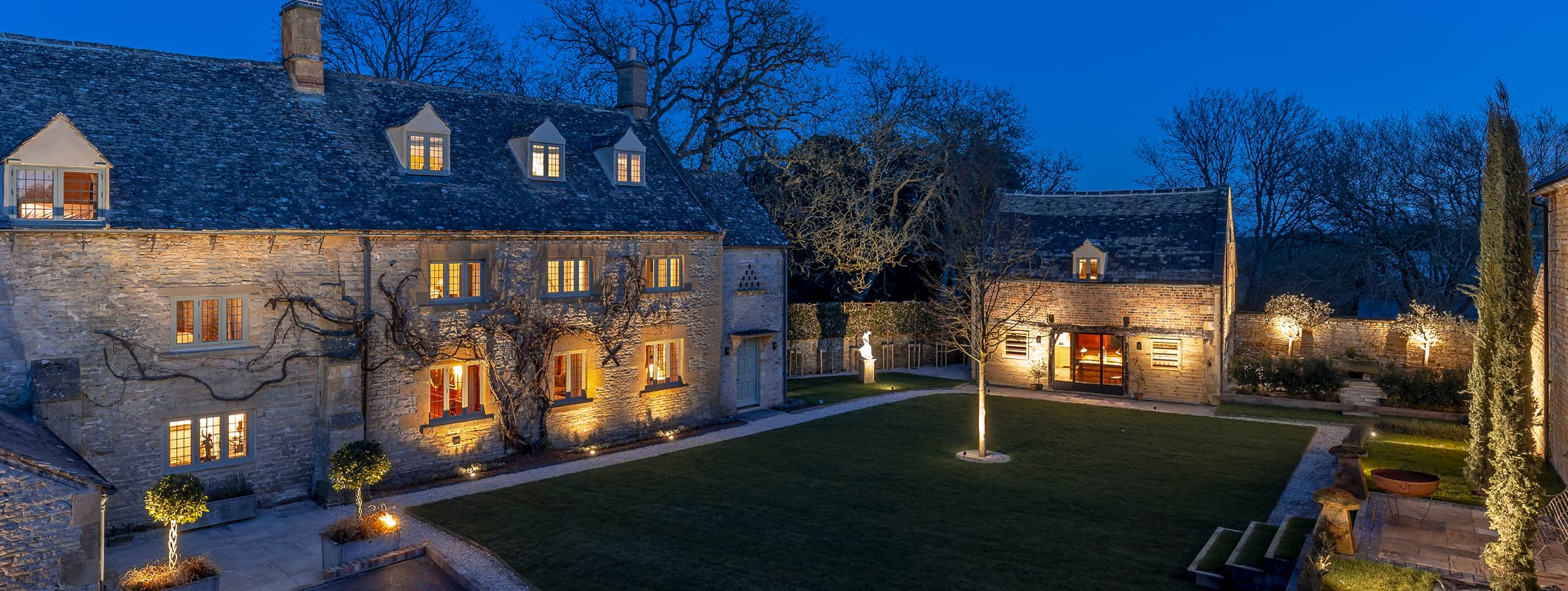 Cotswold stone manor house and courtyard lit up at dusk, with warm glowing windows, manicured lawn, and landscaped gardens