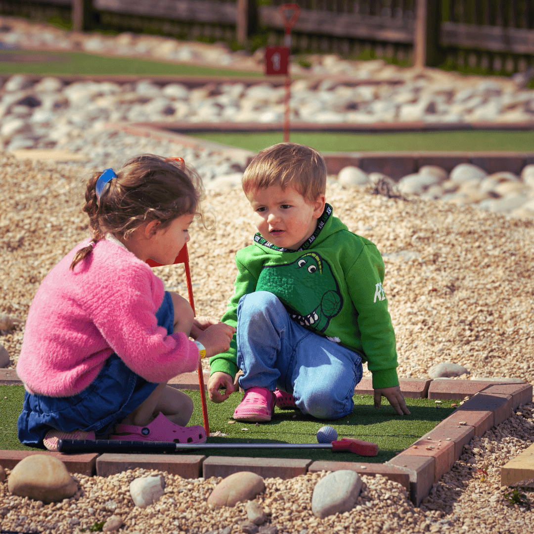 Two young children crouched on a mini golf green, playing together with clubs and a purple ball at a holiday park in the Cot…