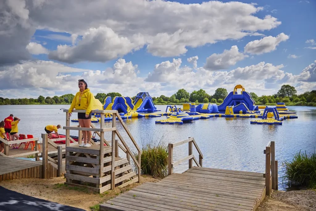 Inflatable aqua park on a calm Cotswolds lake with blue and yellow obstacles, slides, and a wooden jetty entry point on a su…