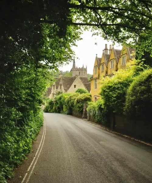 Tree-lined country road winding through honey-coloured Cotswolds stone cottages with traditional chimneys and lush gardens