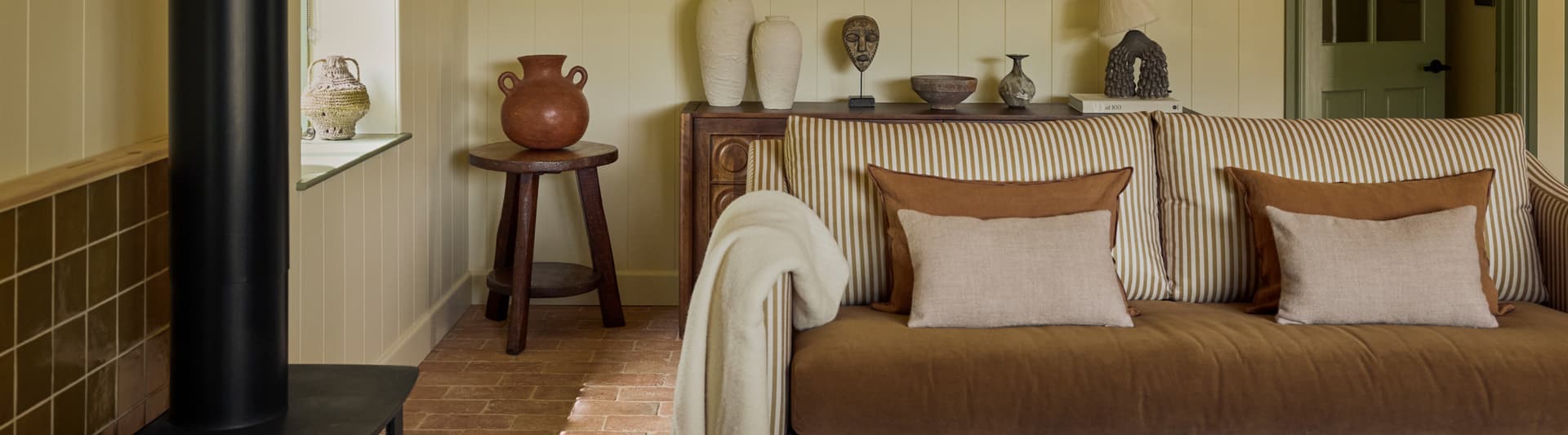 Cosy holiday cottage sitting room with striped sofa, terracotta floor tiles, wood-burning stove and curated ceramic and scul…