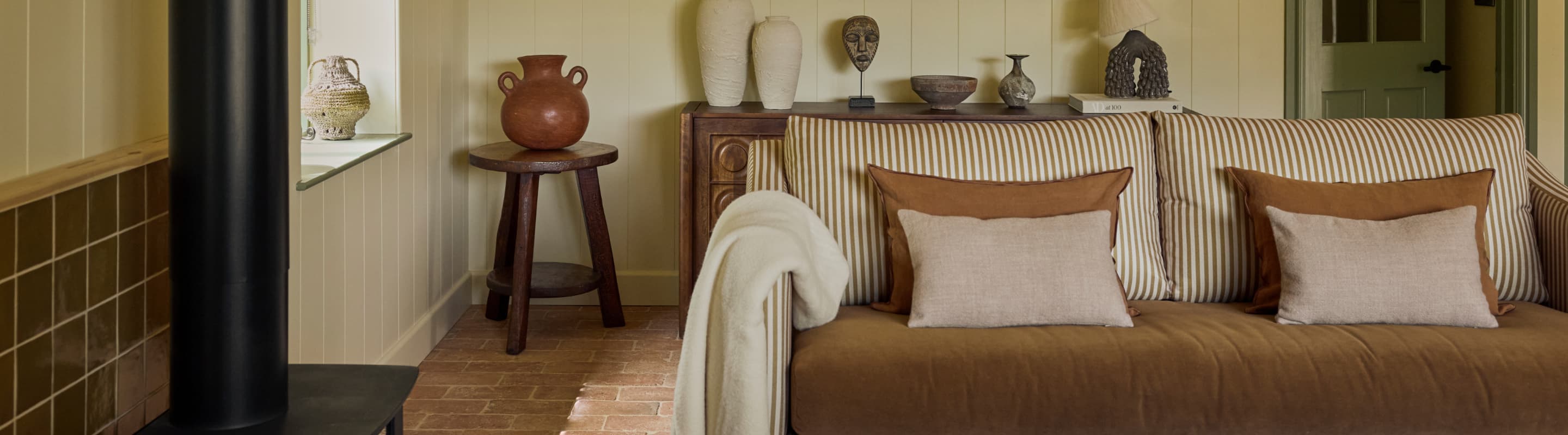 Cosy holiday cottage sitting room with striped sofa, terracotta floor tiles, wood-burning stove and curated ceramic and scul…