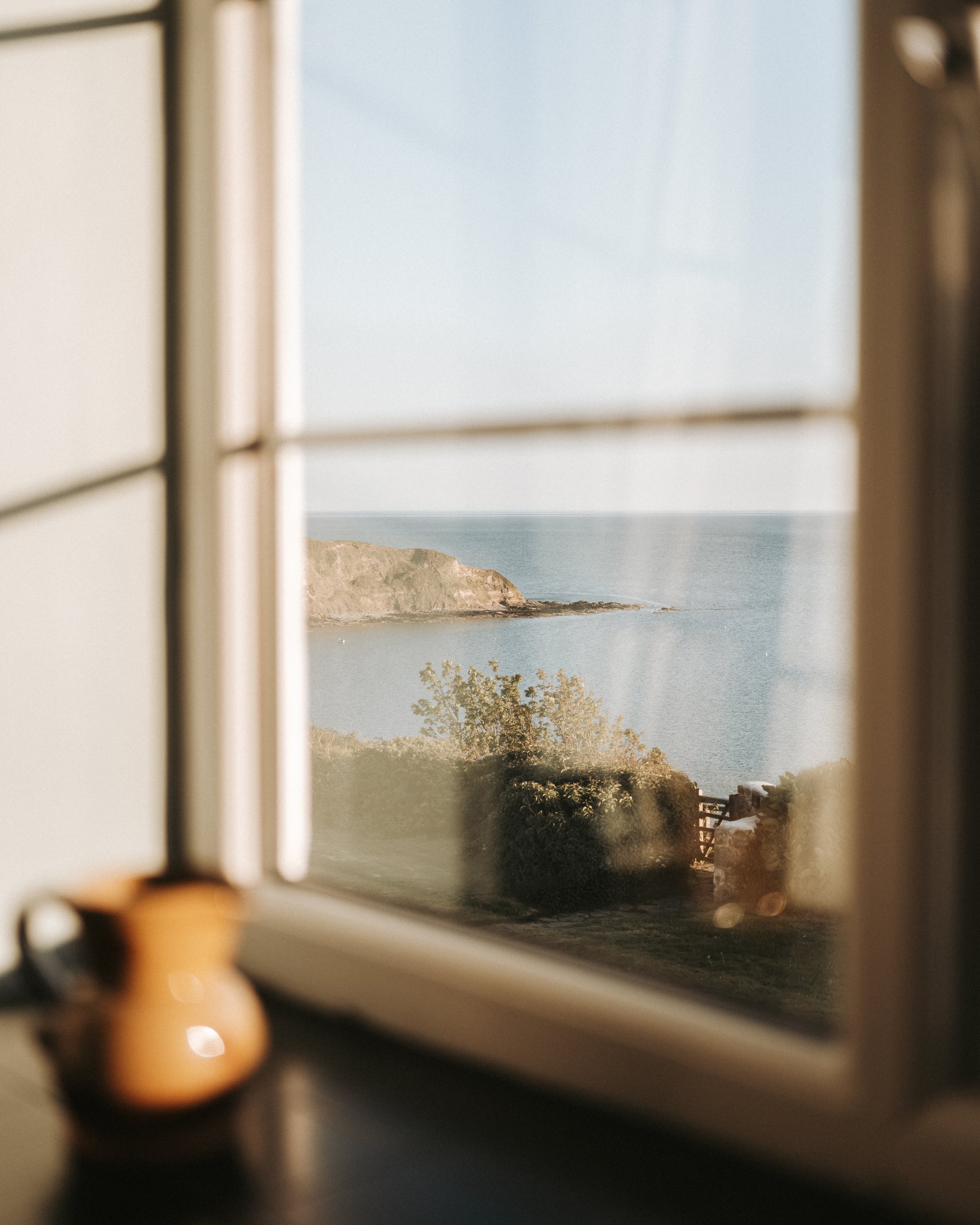 Cottage window framing a sunlit coastal headland and calm sea, with a terracotta jug on the sill in the foreground
