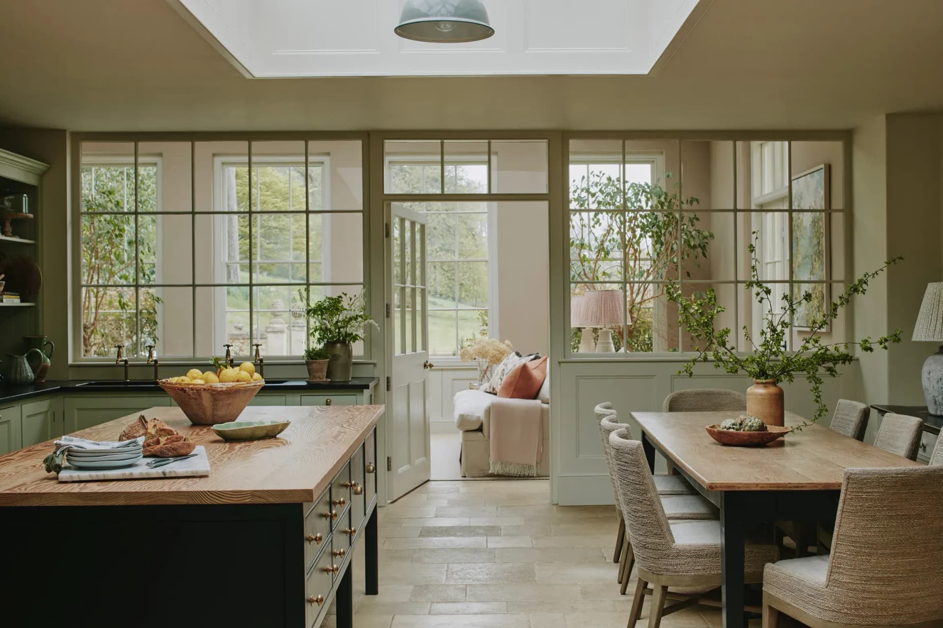 Open-plan kitchen diner in a luxury country cottage with sage green cabinetry, oak island, rooflight, and garden views throu…