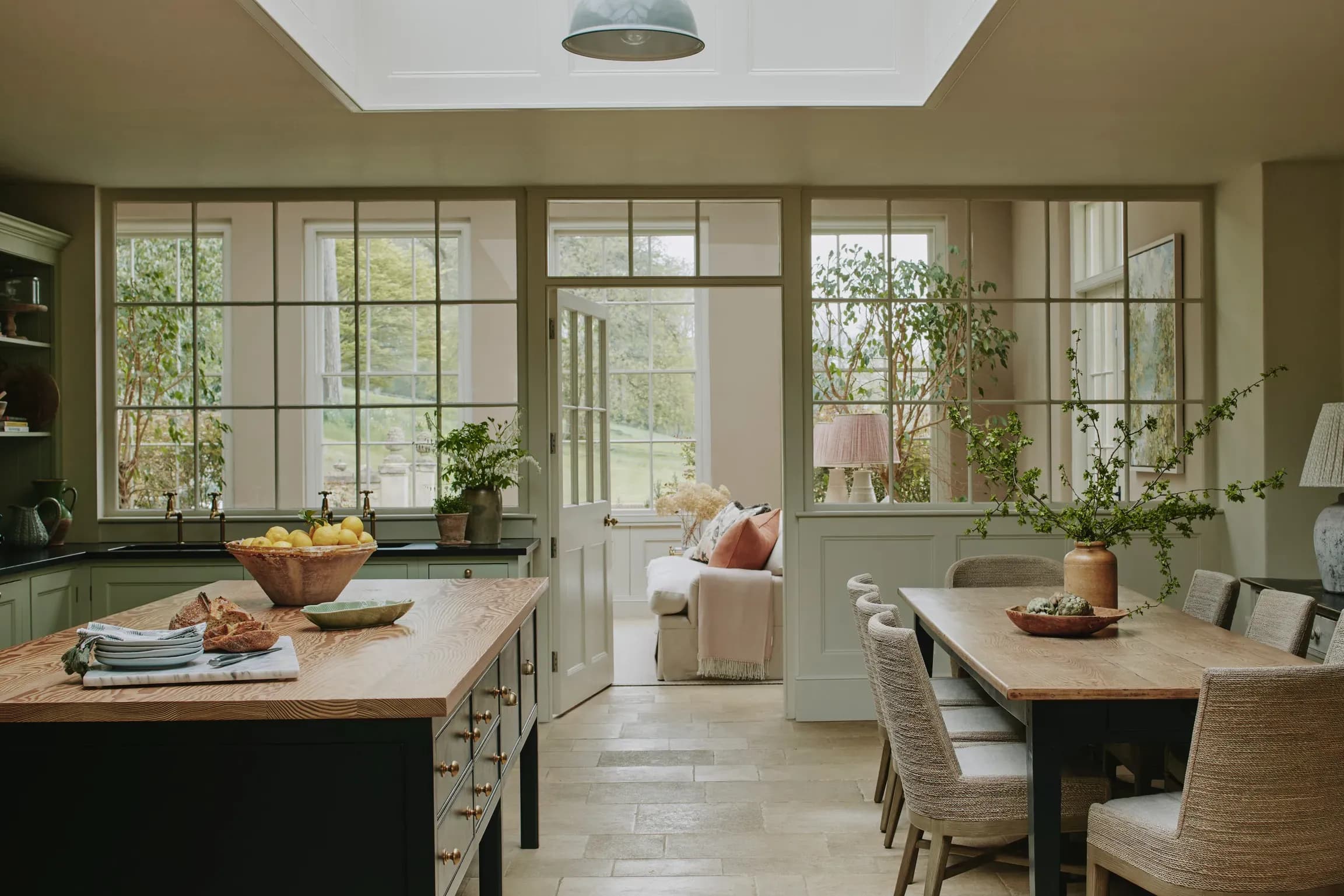 Open-plan kitchen diner in a luxury country cottage with sage green cabinetry, oak island, rooflight, and garden views throu…