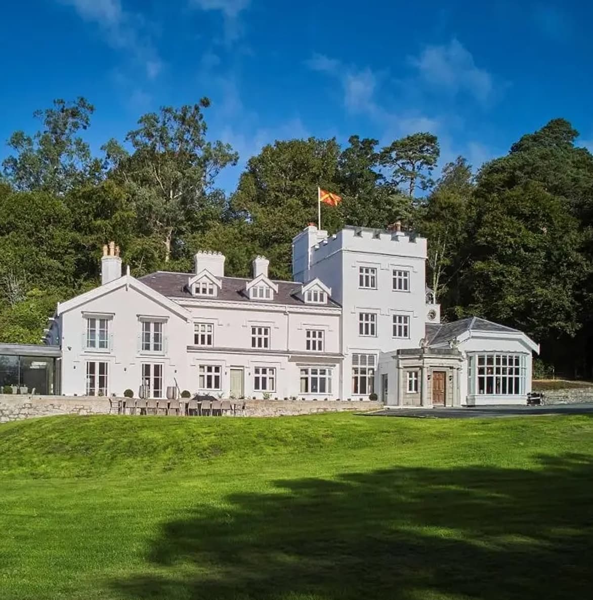 Grand white-rendered country manor with castellated tower, flag flying, orangery entrance, and sweeping lawns set against ma…