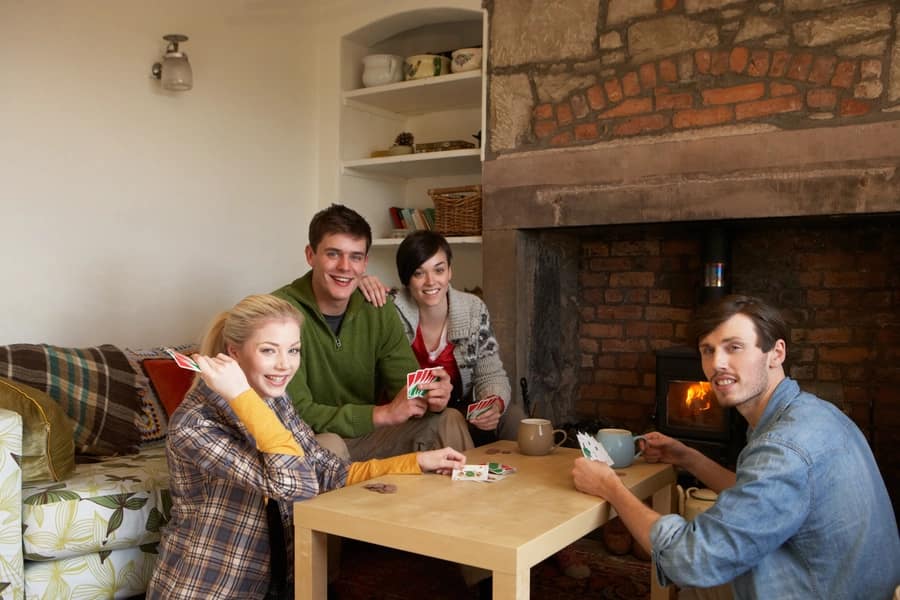 Four friends playing card games around a coffee table in a cosy cottage living room with a log burner glowing in a brick fir…