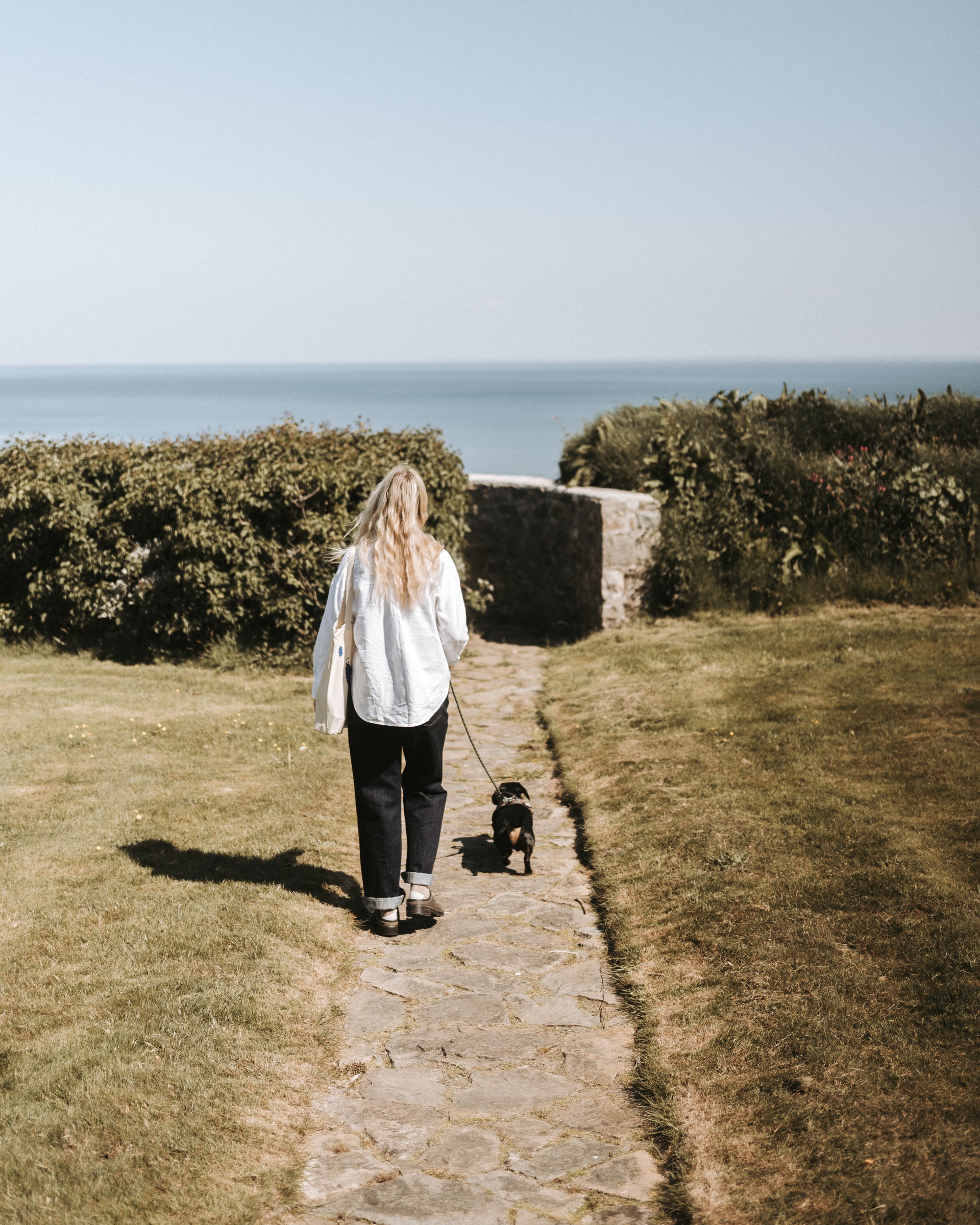 Woman walking a small dog along a stone path towards the sea on a sunny day at a dog-friendly coastal holiday let