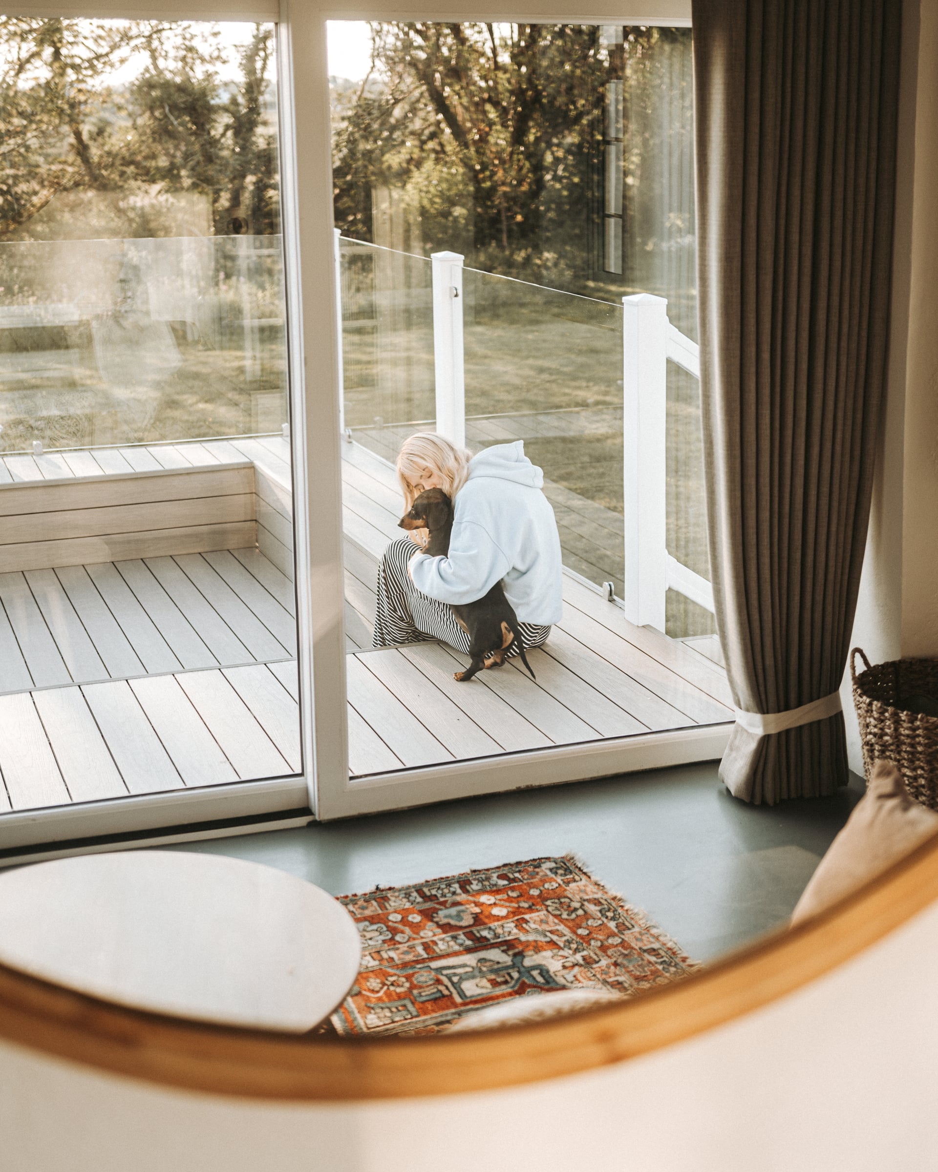 Guest cuddling a dachshund on a timber deck viewed through floor-to-ceiling sliding doors, showing the dog-friendly appeal o…