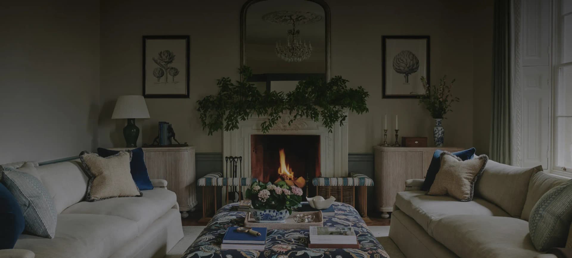 Elegant drawing room with a roaring open fireplace, ornate gilt mirror, botanical prints, and plush sofas styled in blue and…