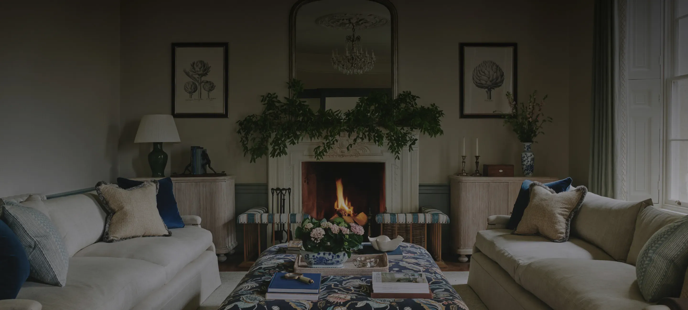 Elegant drawing room with a roaring open fireplace, ornate gilt mirror, botanical prints, and plush sofas styled in blue and…
