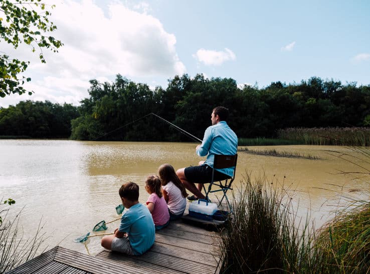 Father and two children fishing from a wooden jetty on a calm lake surrounded by woodland, ideal for family holiday lets