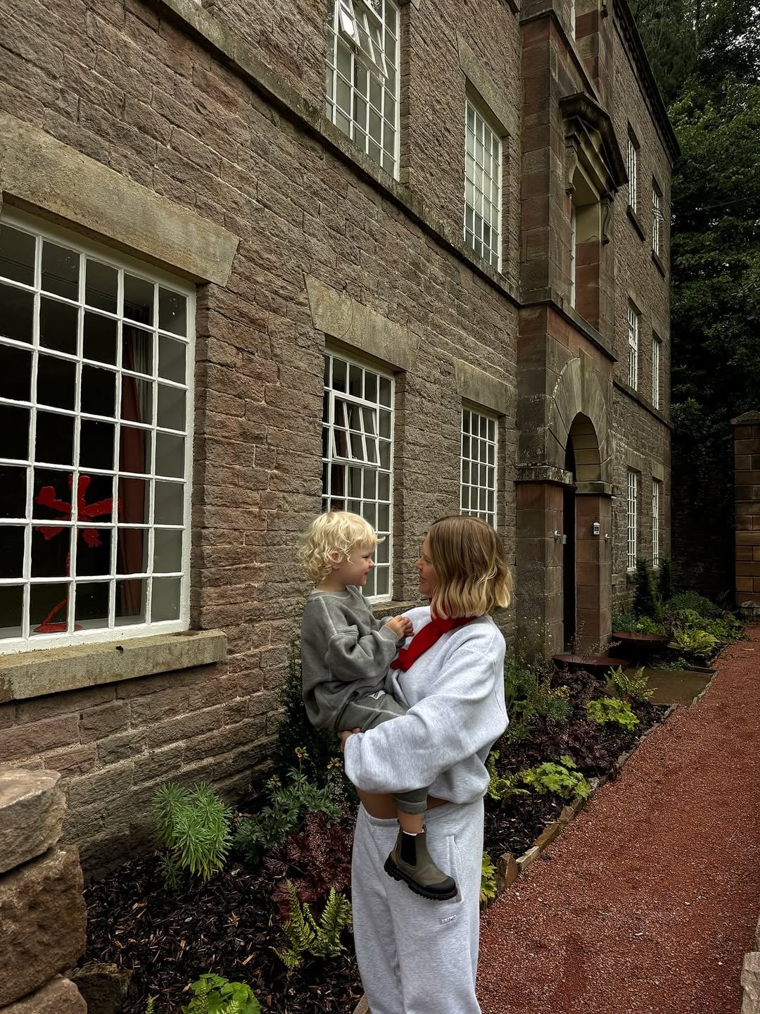 Mother holding a toddler outside a grand stone manor house, ideal for a family holiday let in the UK countryside