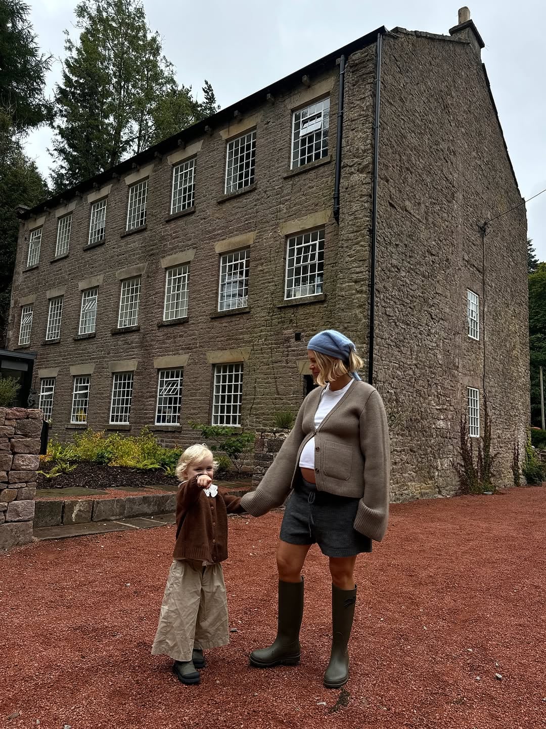 Mother and toddler holding hands outside a grand multi-storey stone mill building converted into a holiday let, surrounded b…