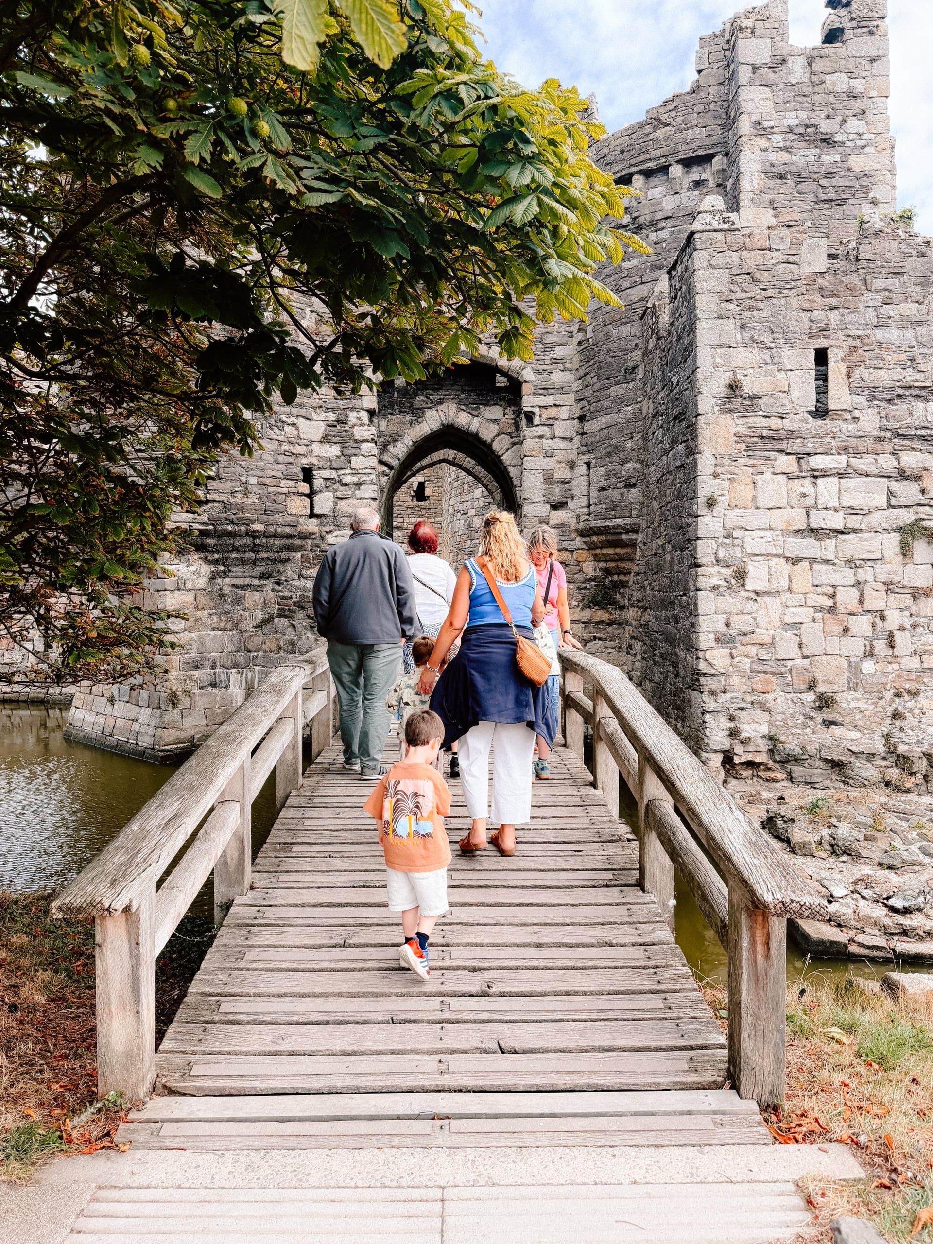 Family crossing a wooden drawbridge into Beaumaris Castle, a medieval Welsh fortress with a moat, on a sunny holiday day out