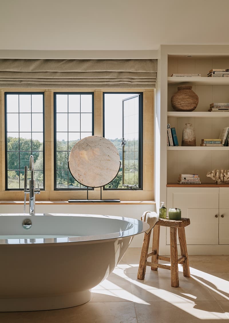 Freestanding oval bath beside leaded windows with countryside views, styled with a rustic wooden stool and built-in shelving…