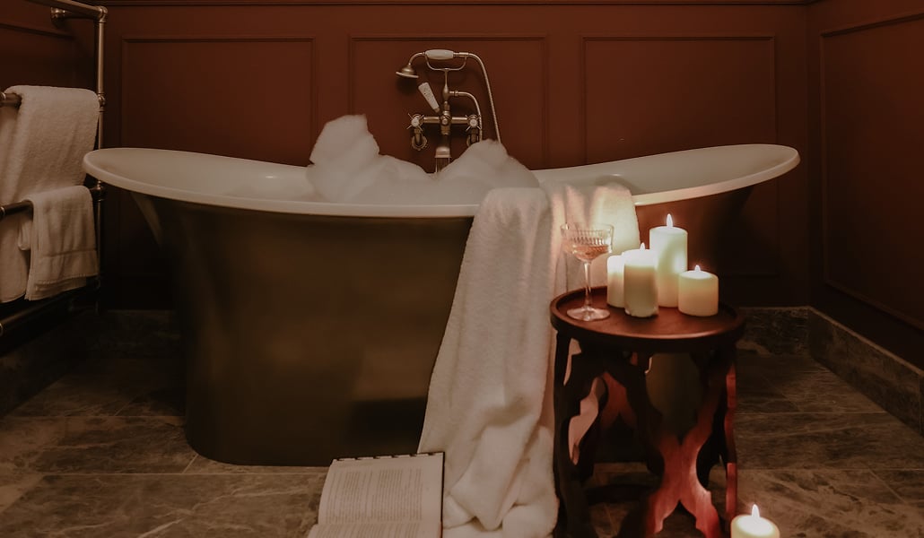 Freestanding roll-top bath filled with bubbles, lit by pillar candles and a champagne glass on a side table, set against dee…