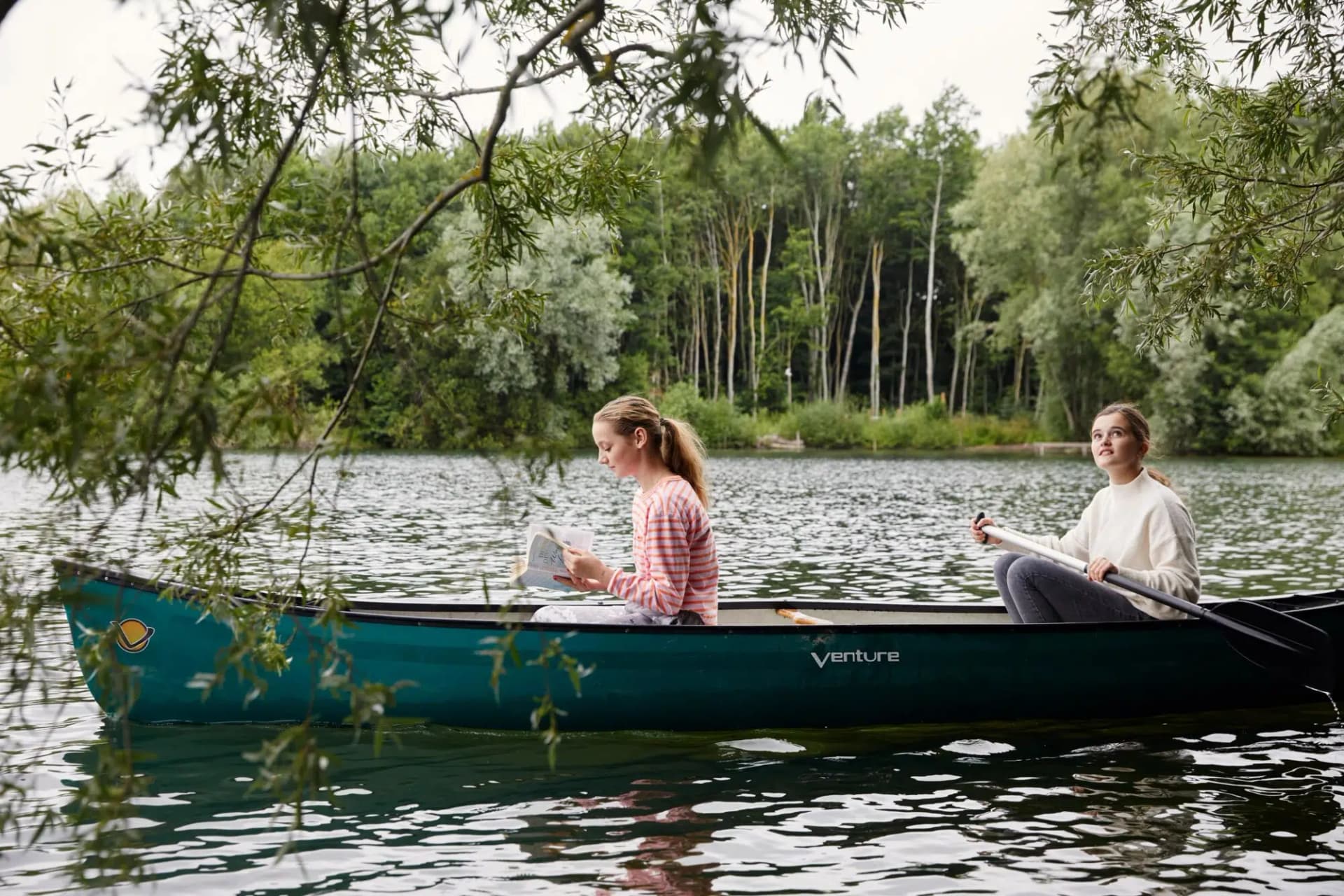 Two girls enjoying a canoe on a calm tree-lined lake, one reading and one paddling during a countryside holiday let stay