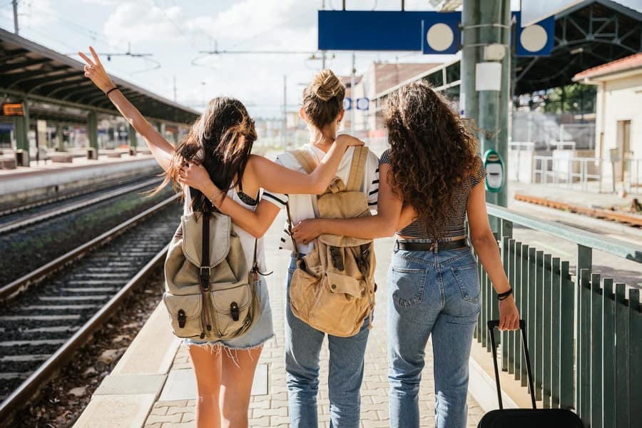 Three friends with backpacks and luggage walking along a sunny train platform, setting off on a group holiday