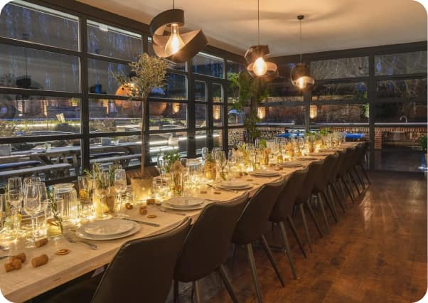 Long dining table set for a large group with glowing candles, wine glasses, and copper pendant lights in a glass-walled dini…