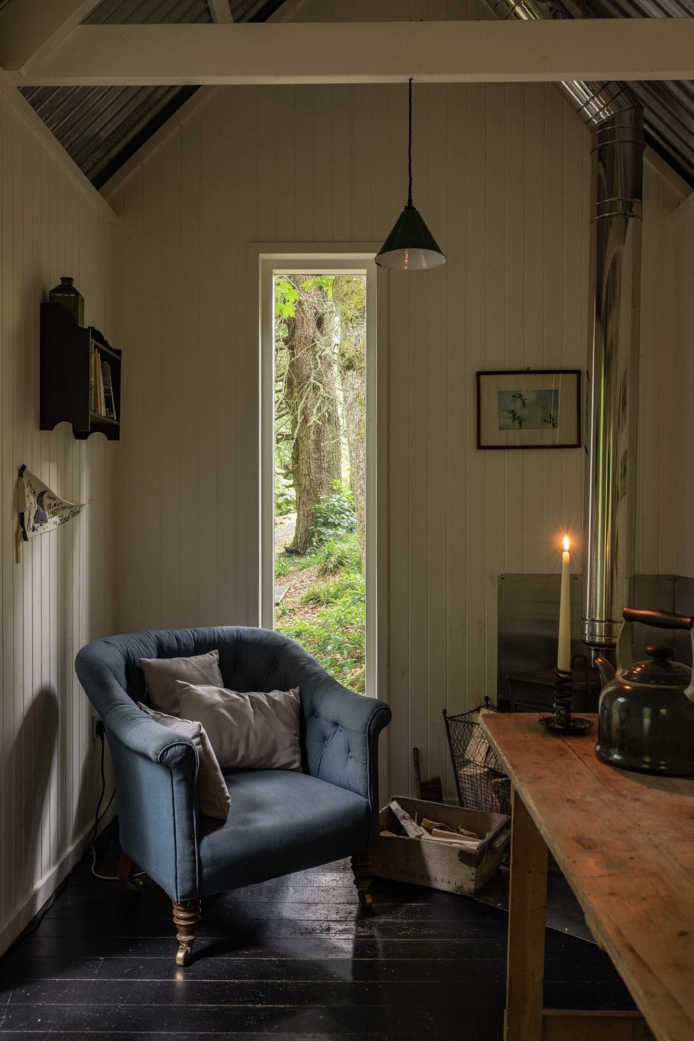 Cosy reading nook in a woodland cabin with a tufted blue armchair, narrow window framing forest trees, lit candle, and log b…