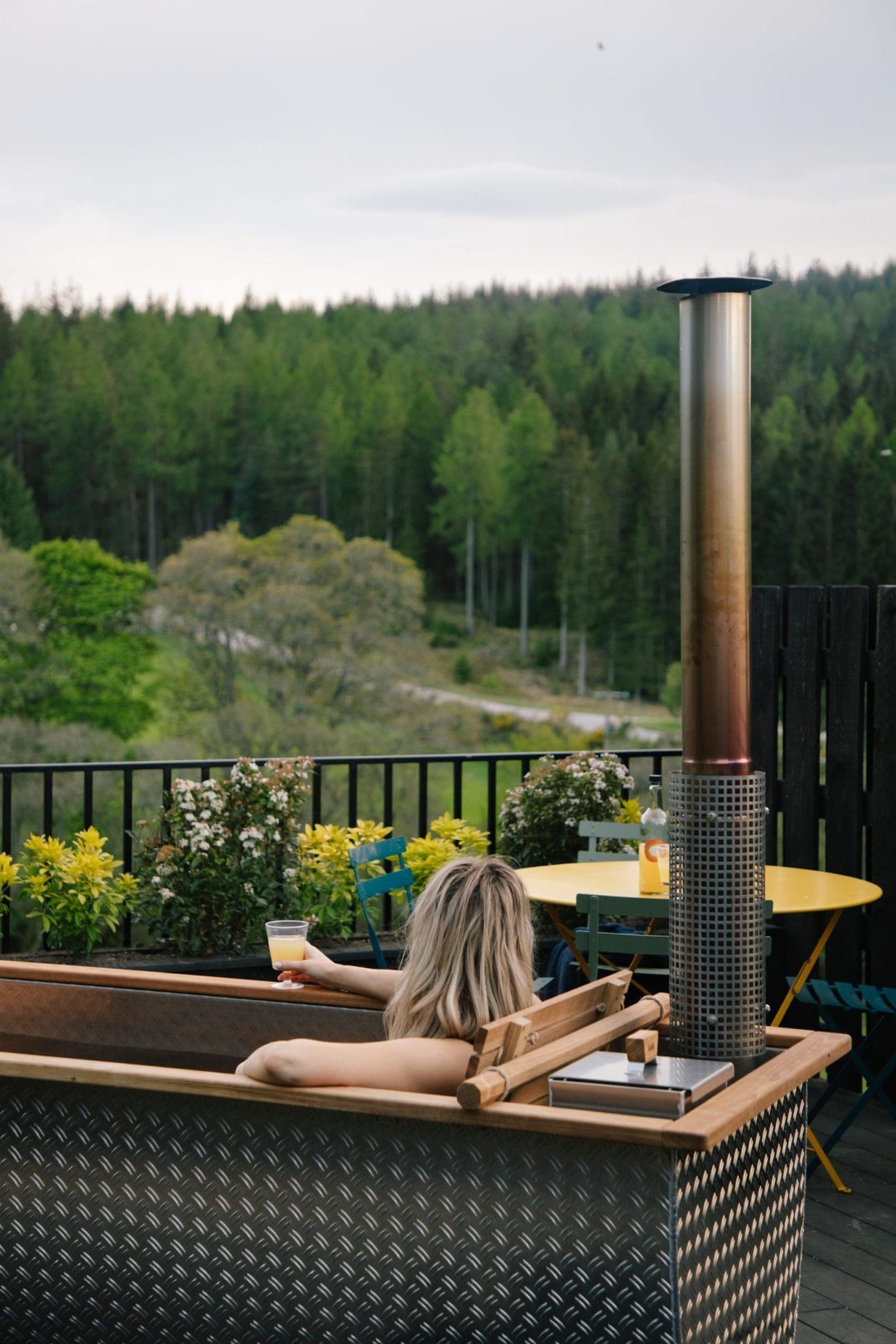 Guest relaxing in a wood-fired hot tub on a decked terrace with a drink, overlooking a lush Scottish forest valley