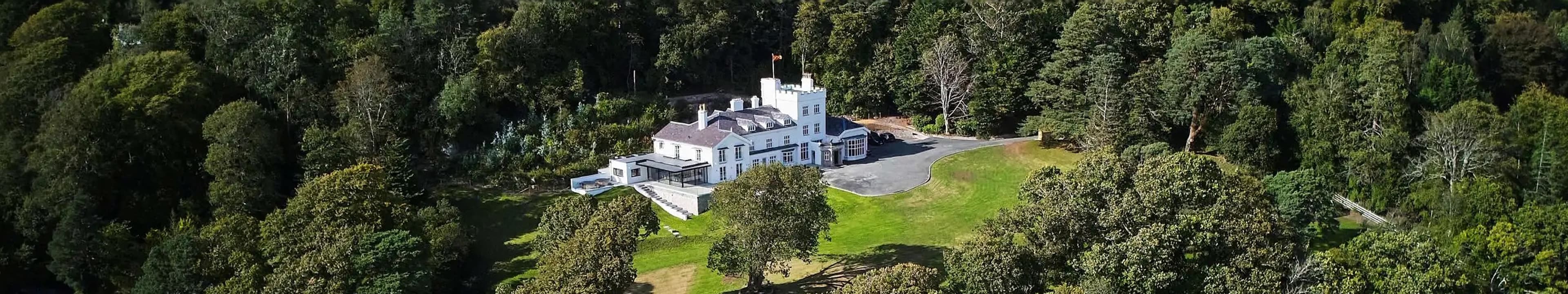 Aerial drone view of a grand white country manor surrounded by dense woodland with sweeping lawns and a private driveway
