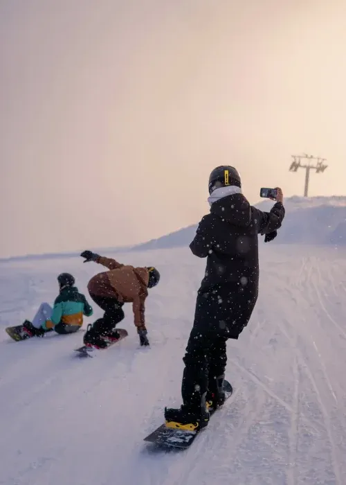 Family snowboarding together on snowy mountain slope with ski lift visible in background during golden hour
