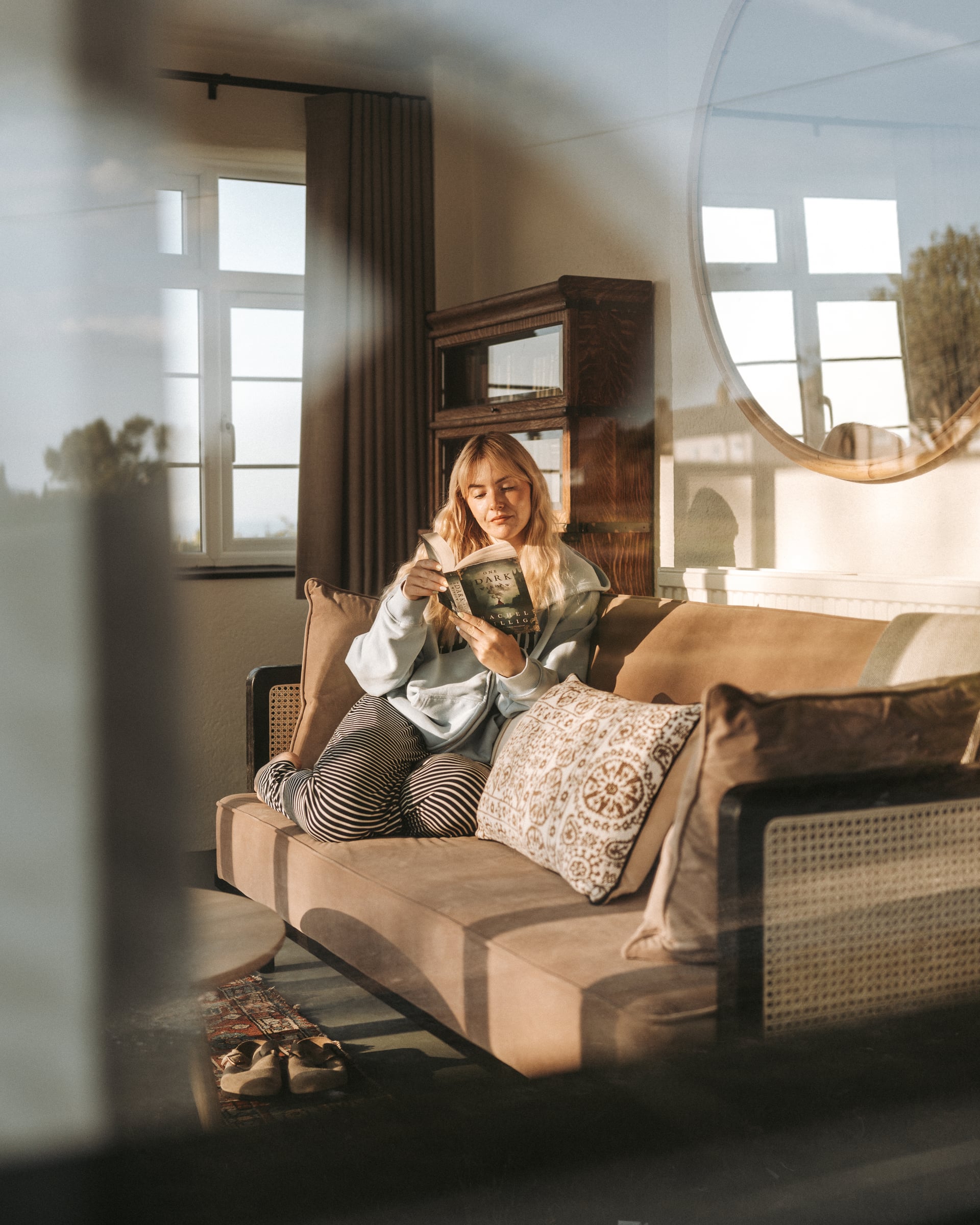 Guest relaxing with a book on a velvet sofa in a sun-drenched holiday let living room with vintage wooden cabinet and round…
