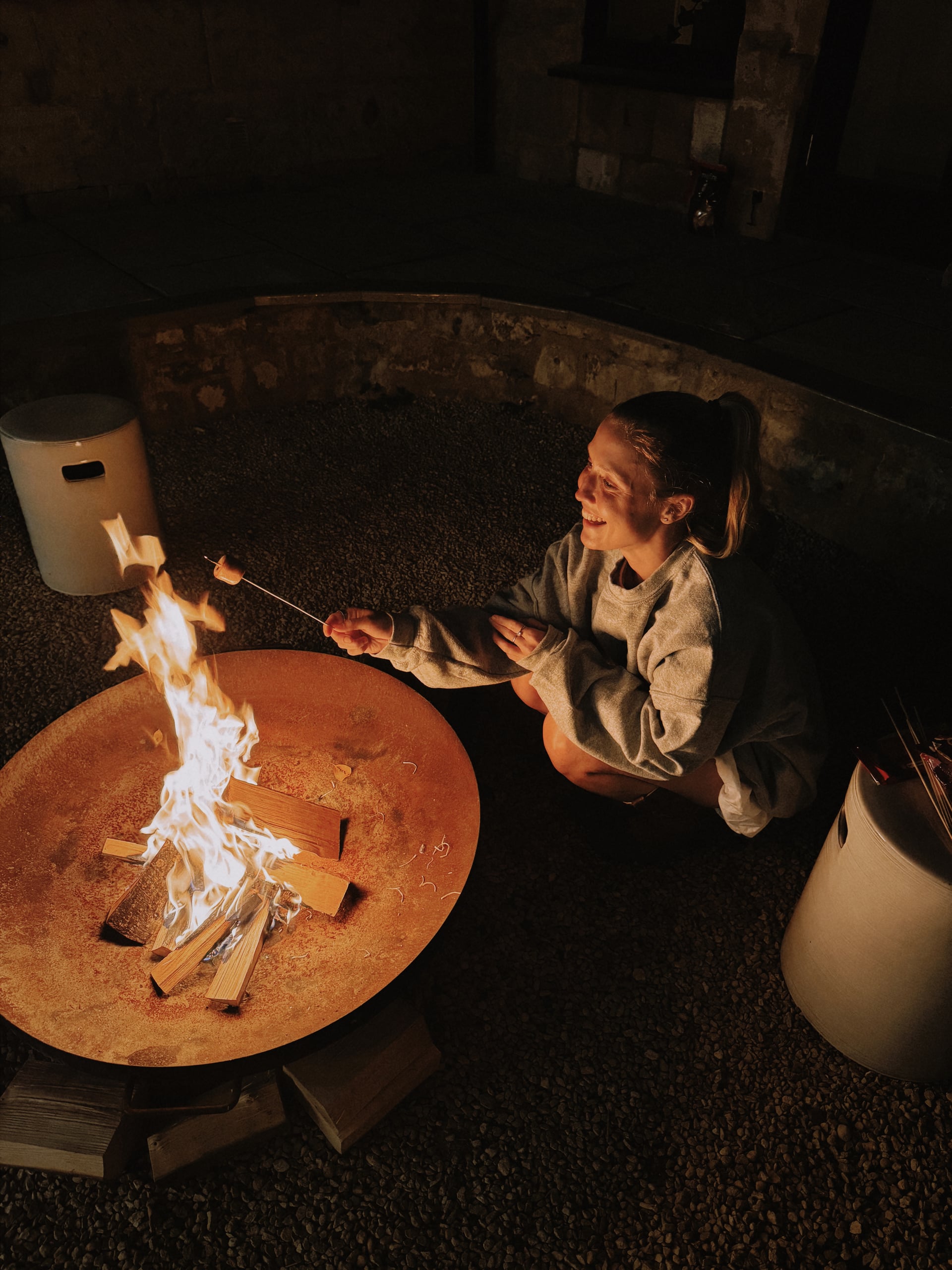 Guest toasting marshmallows over a glowing outdoor fire pit on a gravel terrace at a luxury holiday let