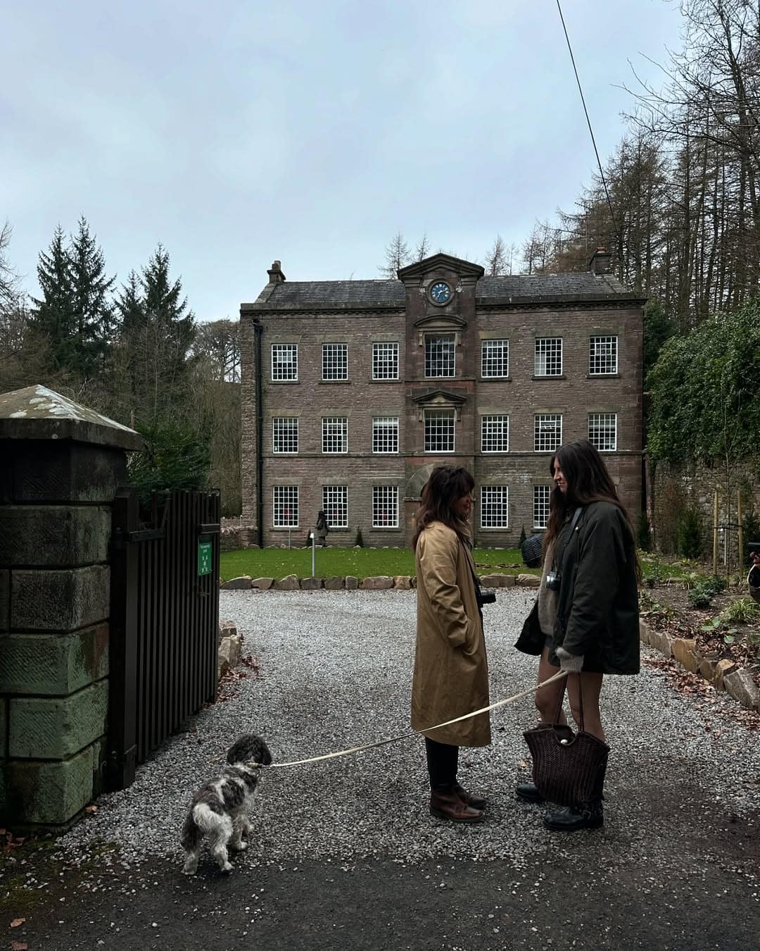 Two women with a small dog on a lead chat on the gravel drive outside a grand Georgian stone manor house surrounded by woodl…