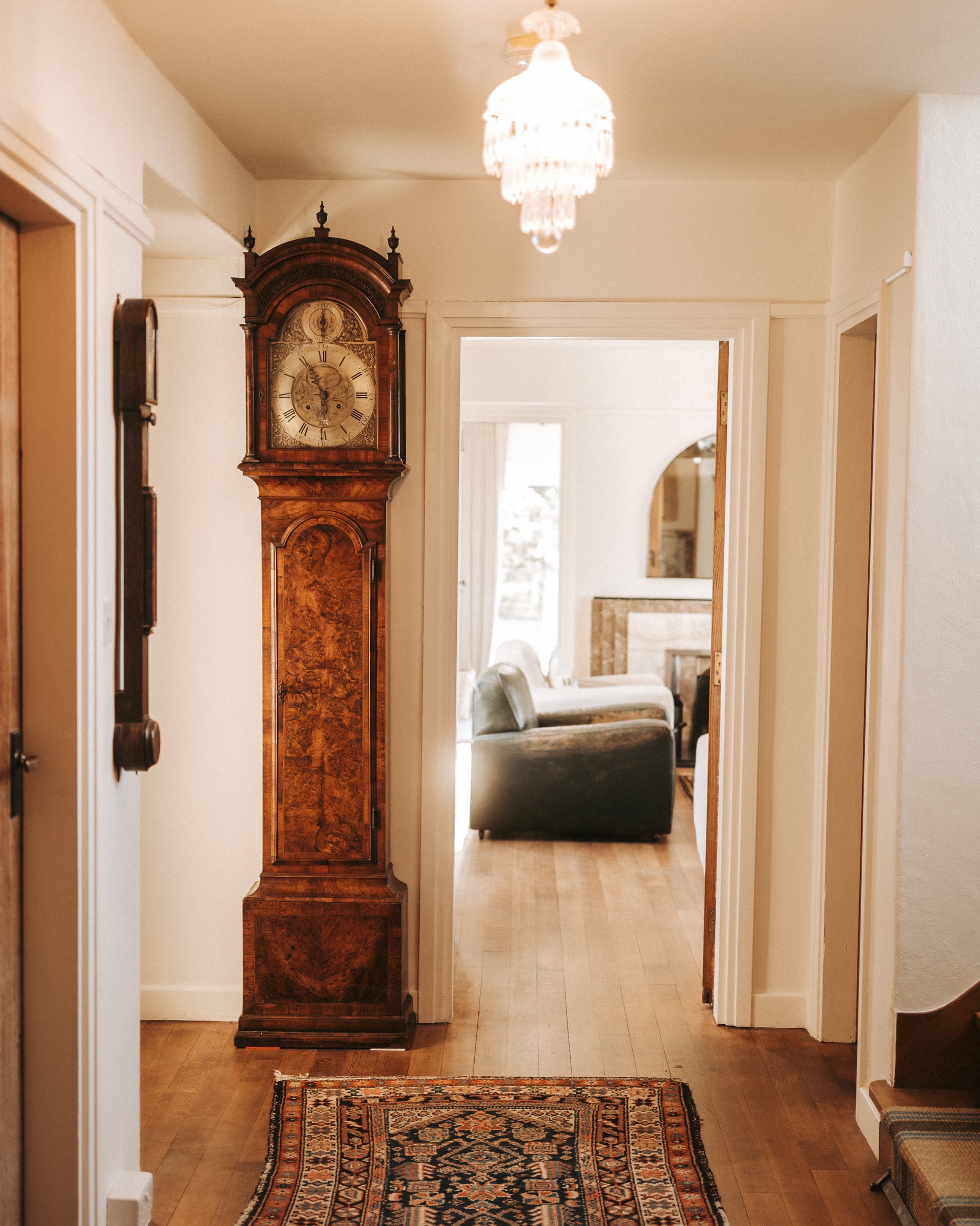 Hallway of a luxury holiday cottage featuring an antique grandfather clock, Persian rug, and crystal chandelier leading to a…