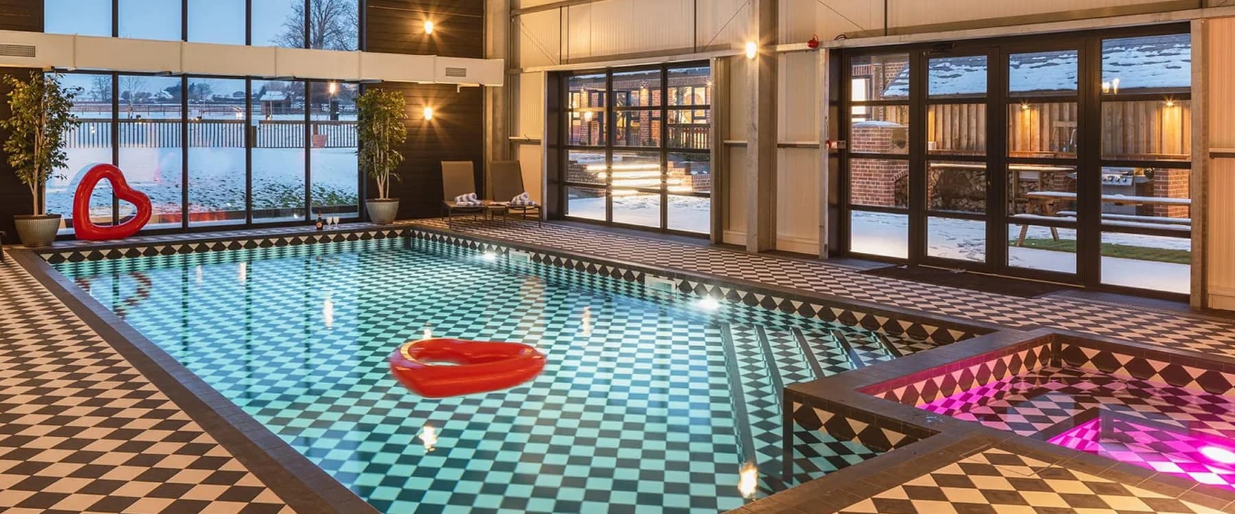 Indoor heated pool with monochrome chequerboard tiles, red heart float, and adjacent hot tub, overlooking a snow-dusted coun…