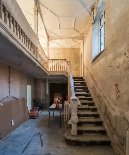 Grand staircase hall mid-renovation in a historic country house, with ornate balustrade, coffered ceiling, and construction…