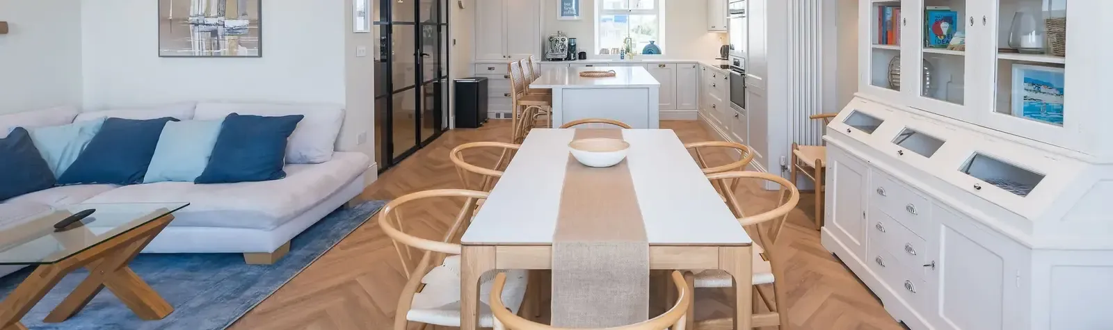Open-plan living, dining and kitchen area with herringbone oak flooring, large white dining table, wishbone chairs, and coas…
