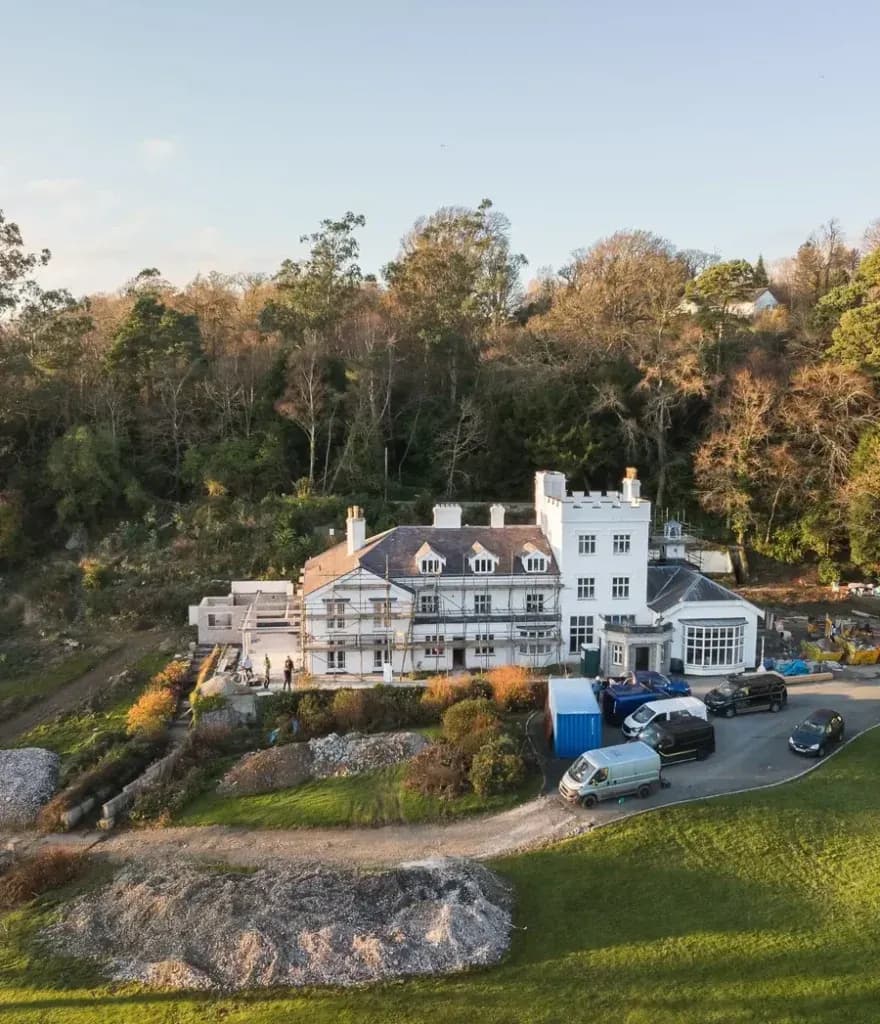 Aerial view of a grand white country house under renovation, surrounded by woodland and lawns in the UK countryside