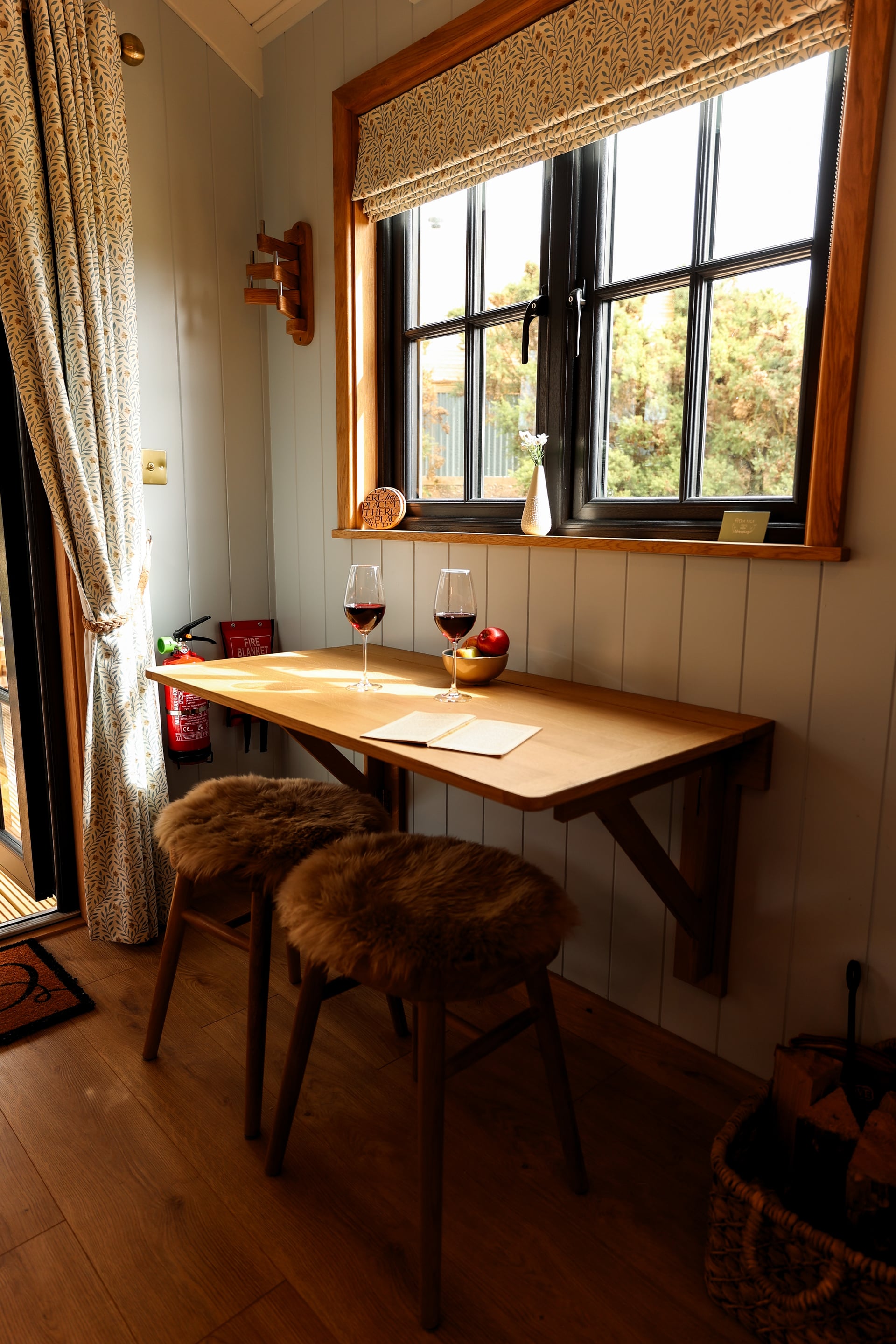 Cosy fold-down dining nook in a holiday let with two glasses of red wine, sheepskin stools, and a black-framed window overlo…