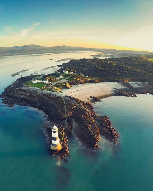 Aerial view of a striped lighthouse on a rocky headland at golden hour, with turquoise waters, a sandy cove, and mountain ba…