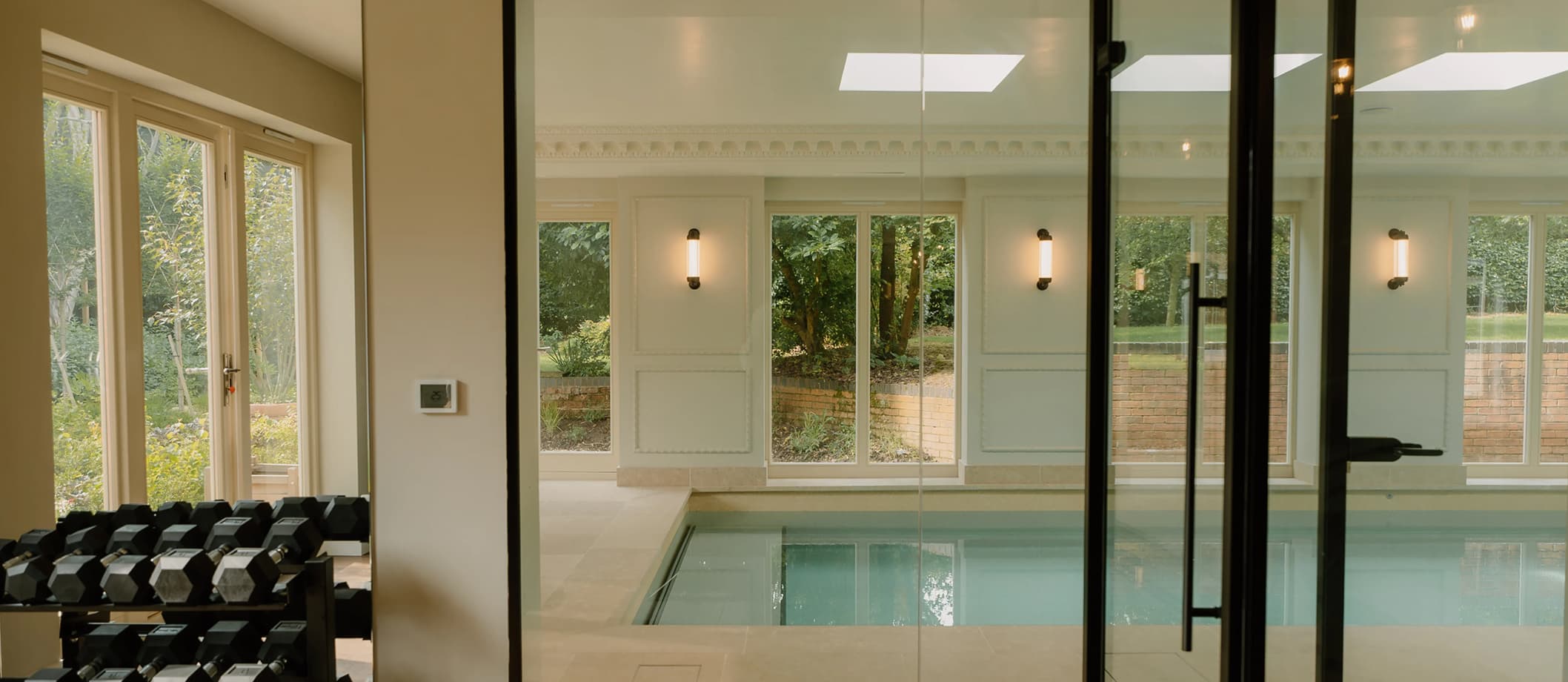 Glass-partitioned indoor swimming pool and gym with dumbbell rack, stone surround, wall sconces, and garden views beyond