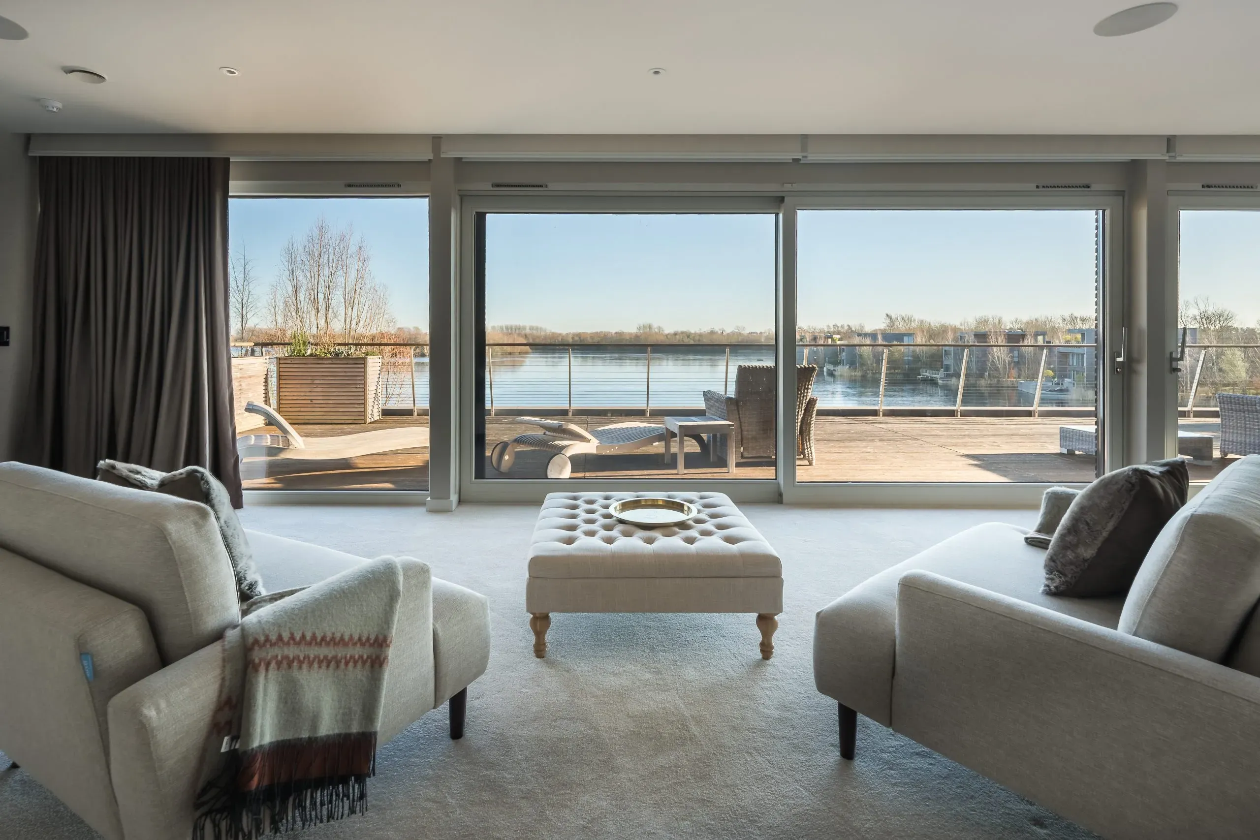 Bright living room with floor-to-ceiling sliding doors opening onto a lakeside terrace with sun loungers and calm water views