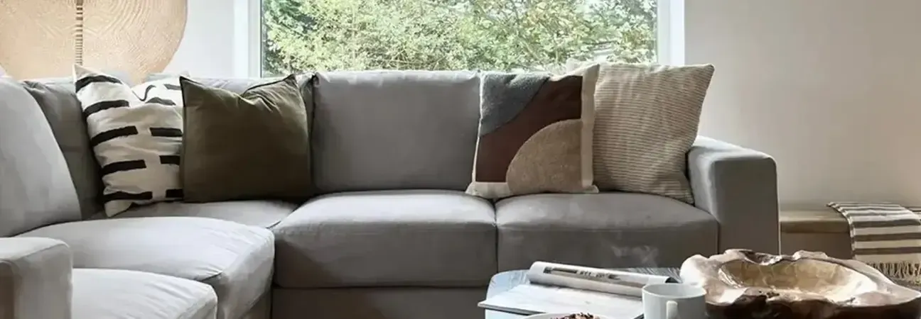 Spacious grey corner sofa with layered cushions in a light-filled holiday cottage living room overlooking woodland