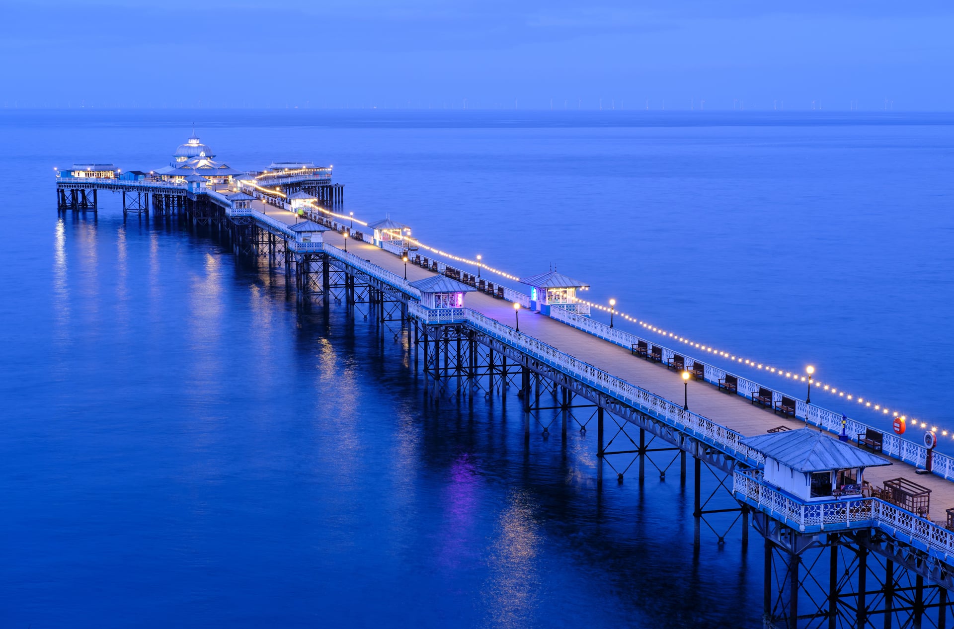 Llandudno Pier illuminated at dusk, its string lights reflecting on the calm blue sea of North Wales