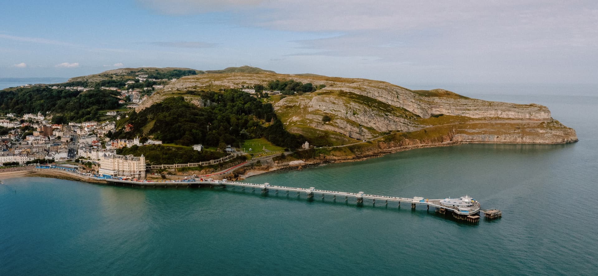 Aerial view of Llandudno pier stretching into calm blue sea, with the Great Orme headland and Victorian town beyond