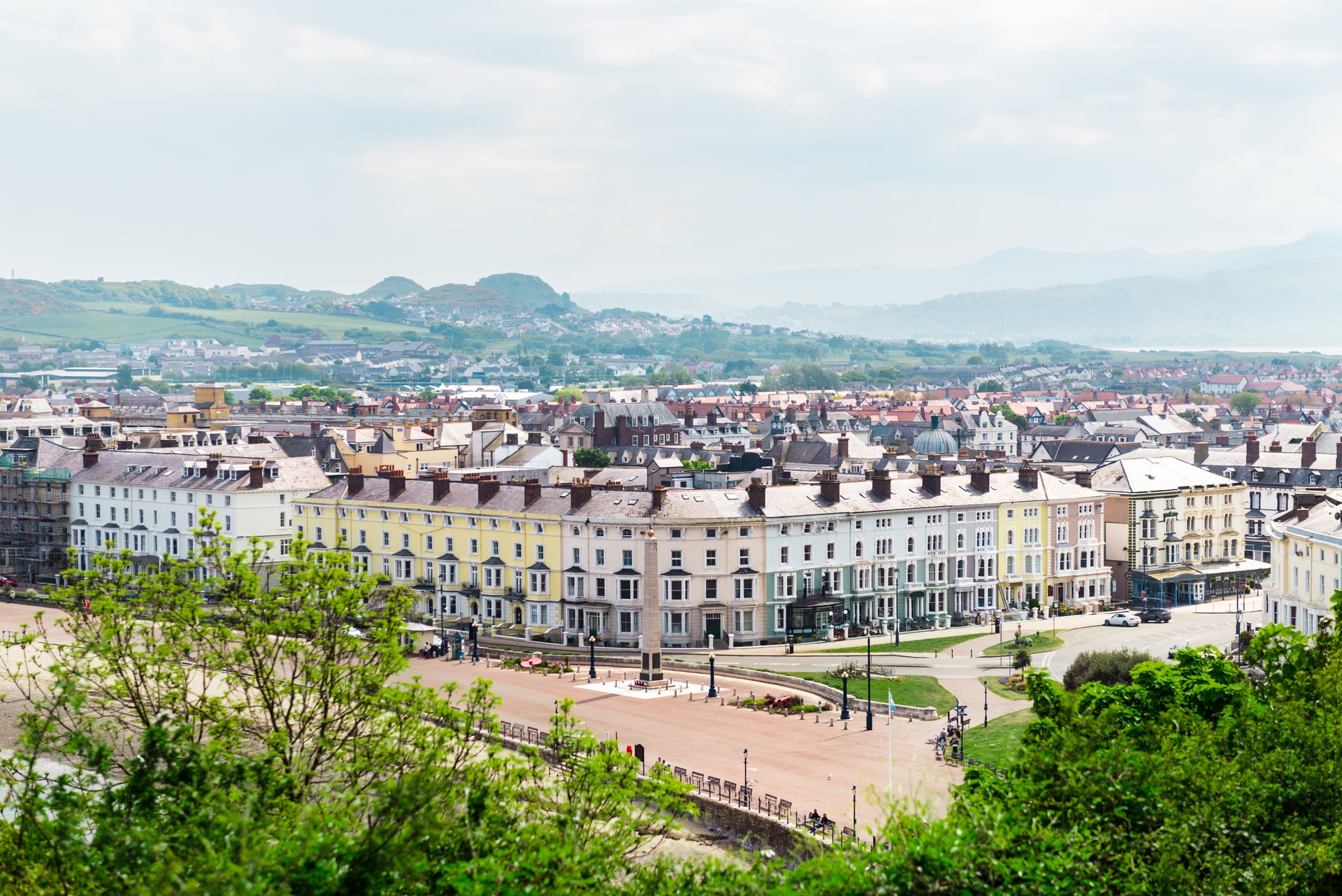 Elevated view over Llandudno promenade with pastel Victorian terraces, a central monument, and misty Welsh hills beyond