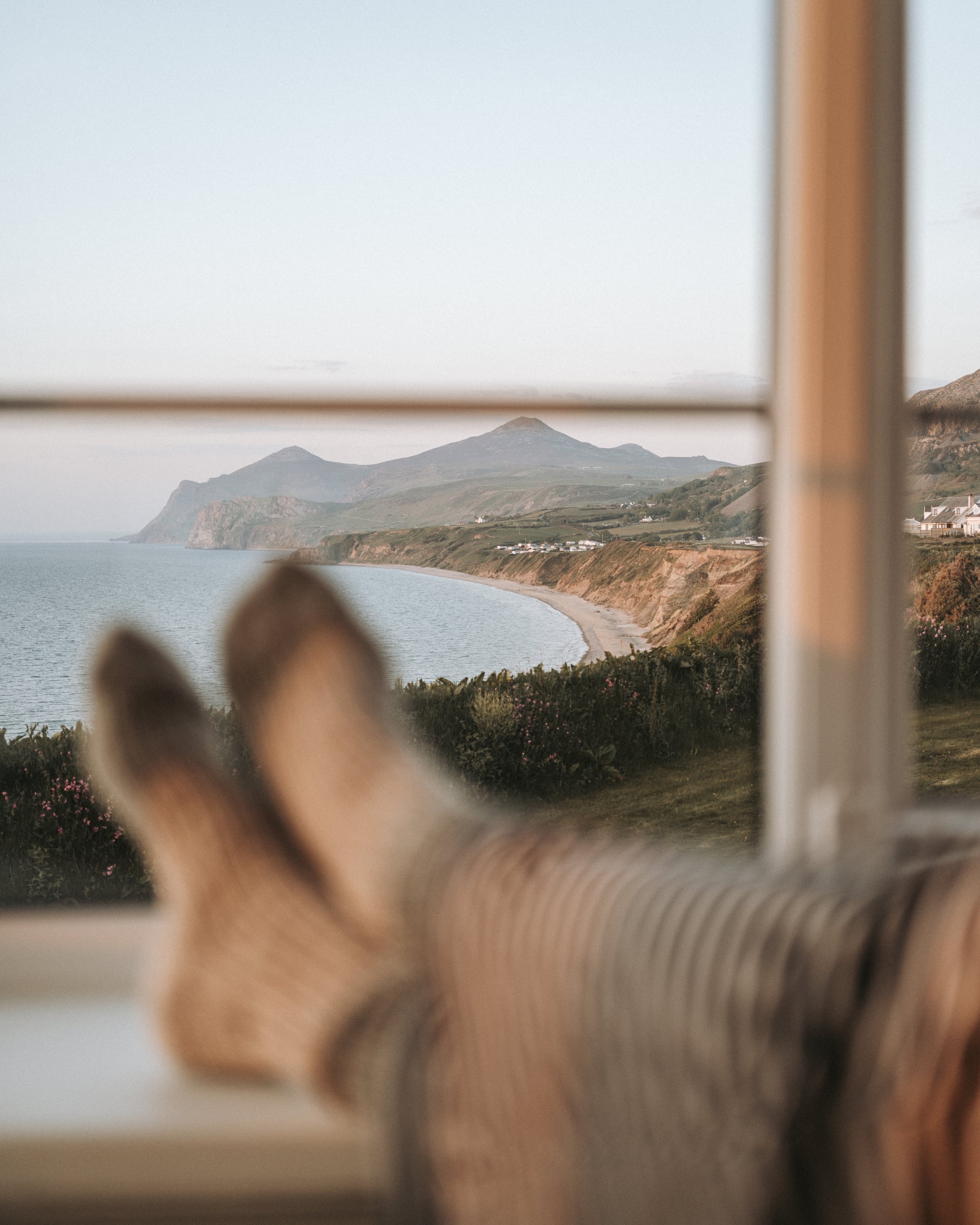 Guest relaxing with feet up enjoying a sweeping window view of a curved sandy bay, coastal cliffs, and mountain on the Llŷn…