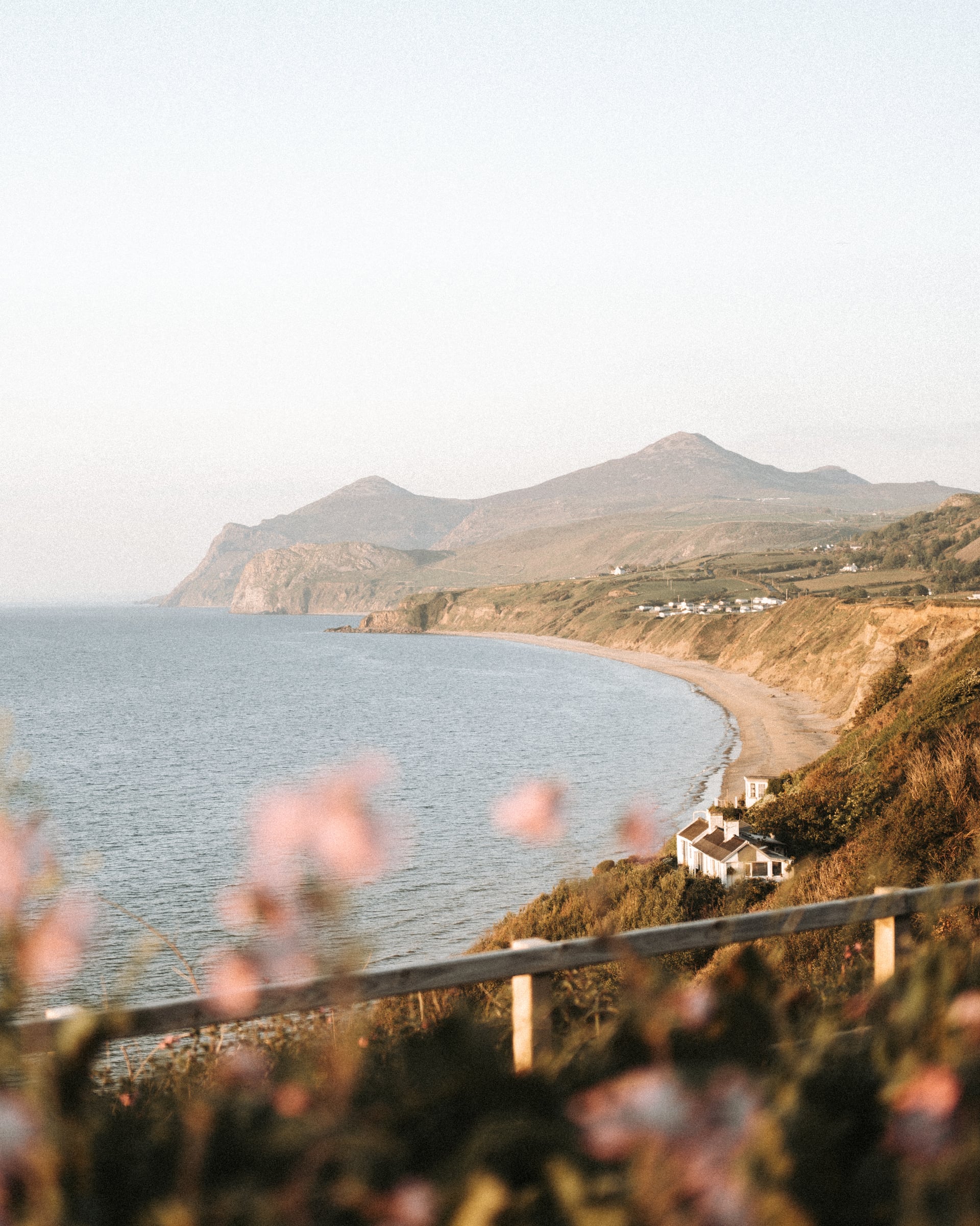 Sweeping bay on the Llŷn Peninsula at golden hour, with a sandy beach curving beneath golden cliffs, a white cottage nestled…