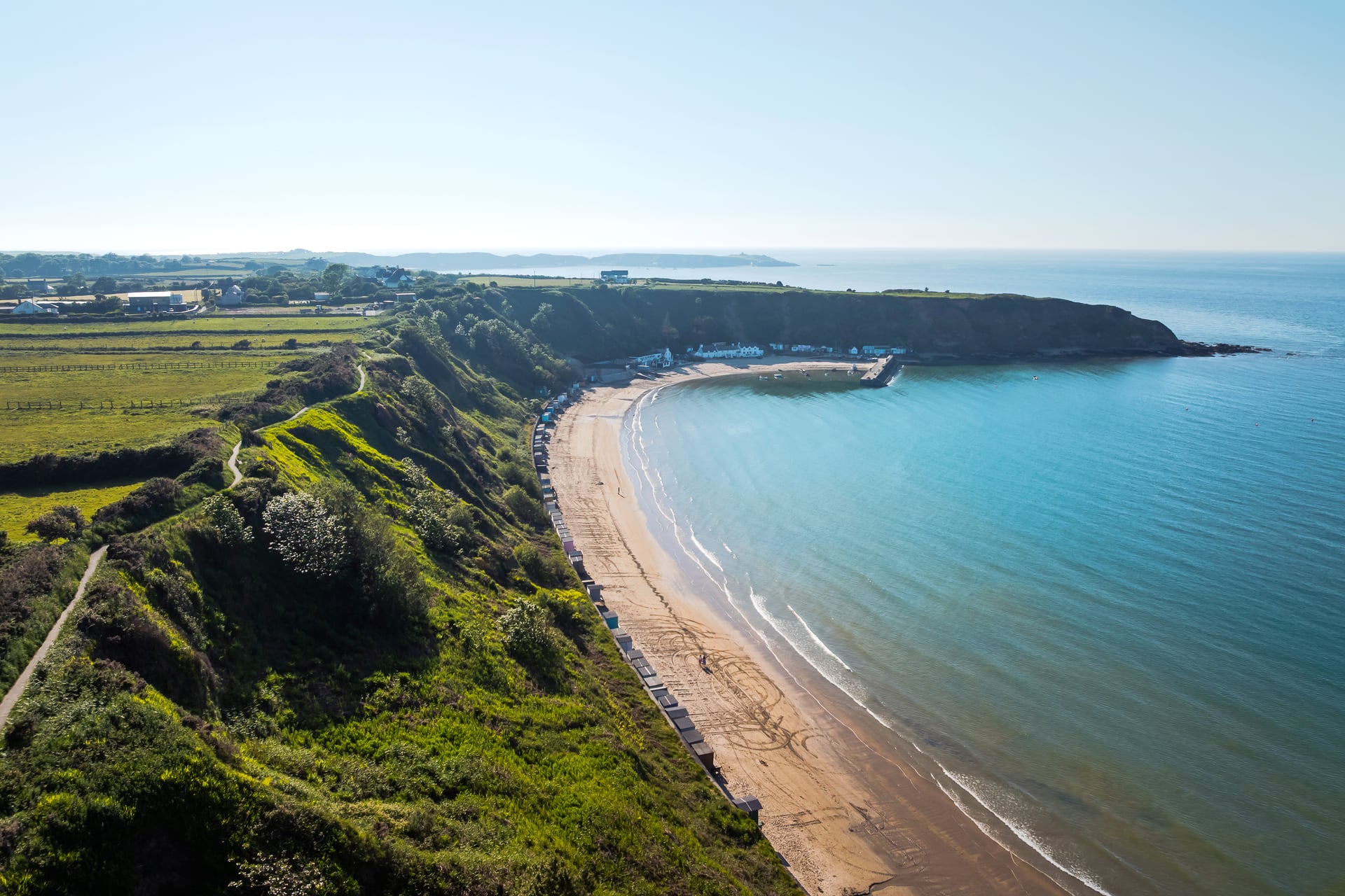 Aerial view of a sweeping sandy bay on the Llŷn Peninsula with colourful beach huts lining green cliffs and calm turquoise s…